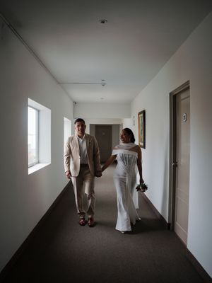 Bride in white gown and groom in beige suit walk hand in hand down a hotel corridor.