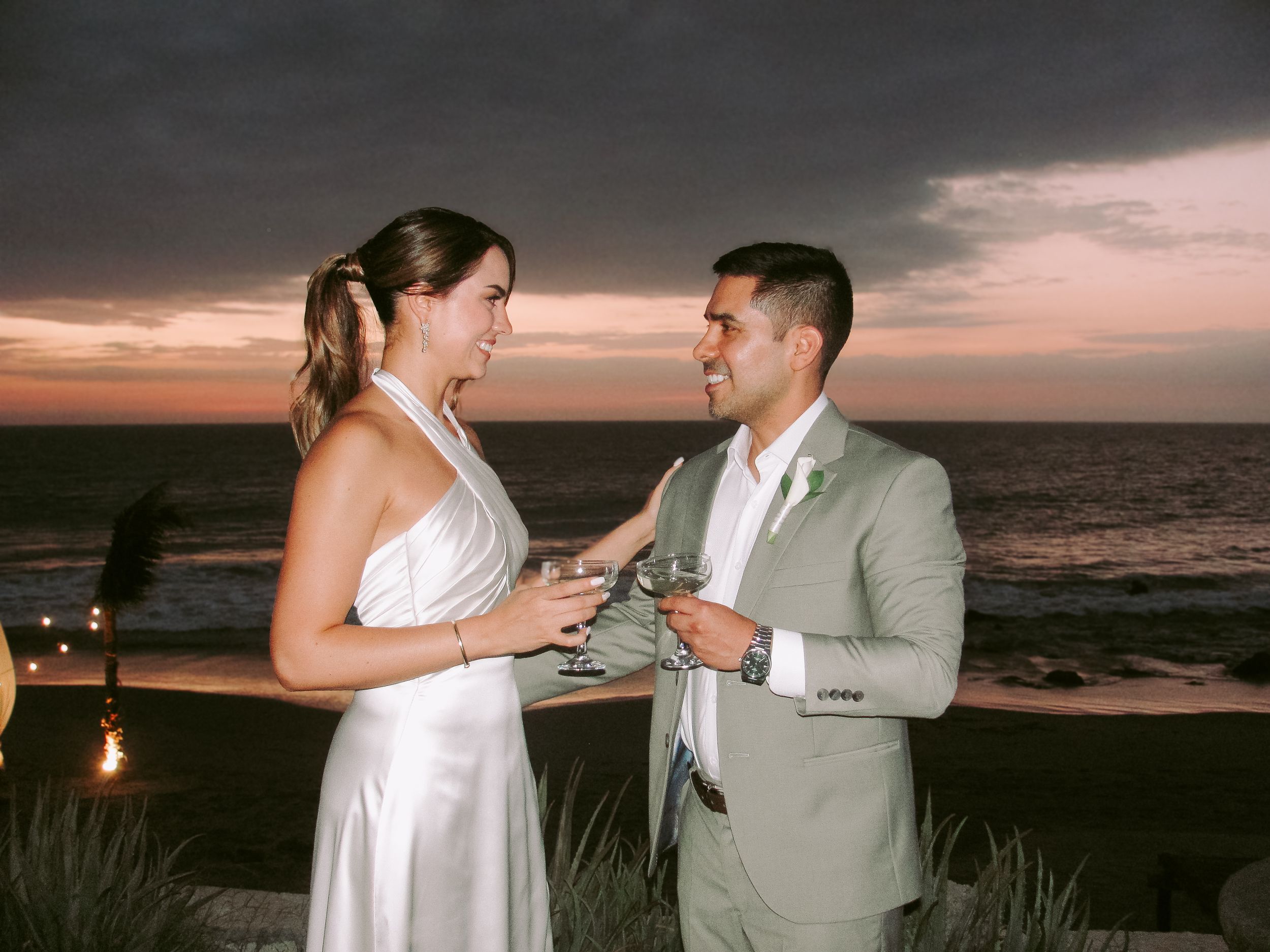 Couple in wedding attire toasting with wine glasses at sunset beach ceremony, bride in white gown, groom in sage suit.