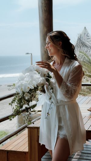 Smiling bride in white lace robe holds orchid bouquet on wooden deck with ocean view.