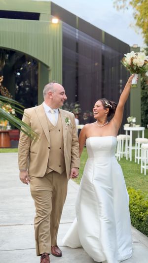 Joyful newlyweds walk together, bride in white strapless gown raises bouquet, groom in tan suit, outdoor wedding venue.
