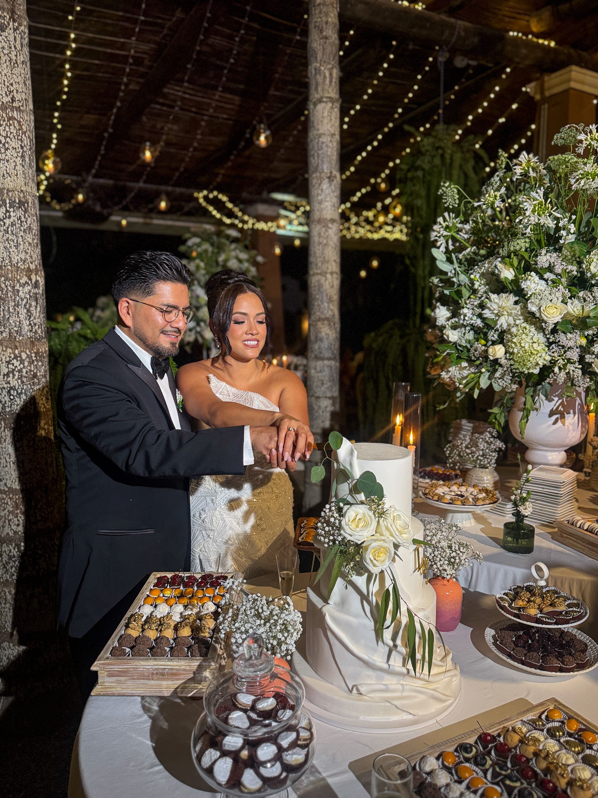 Bride and groom cutting a white wedding cake at an elegant outdoor reception with string lights and floral arrangements.