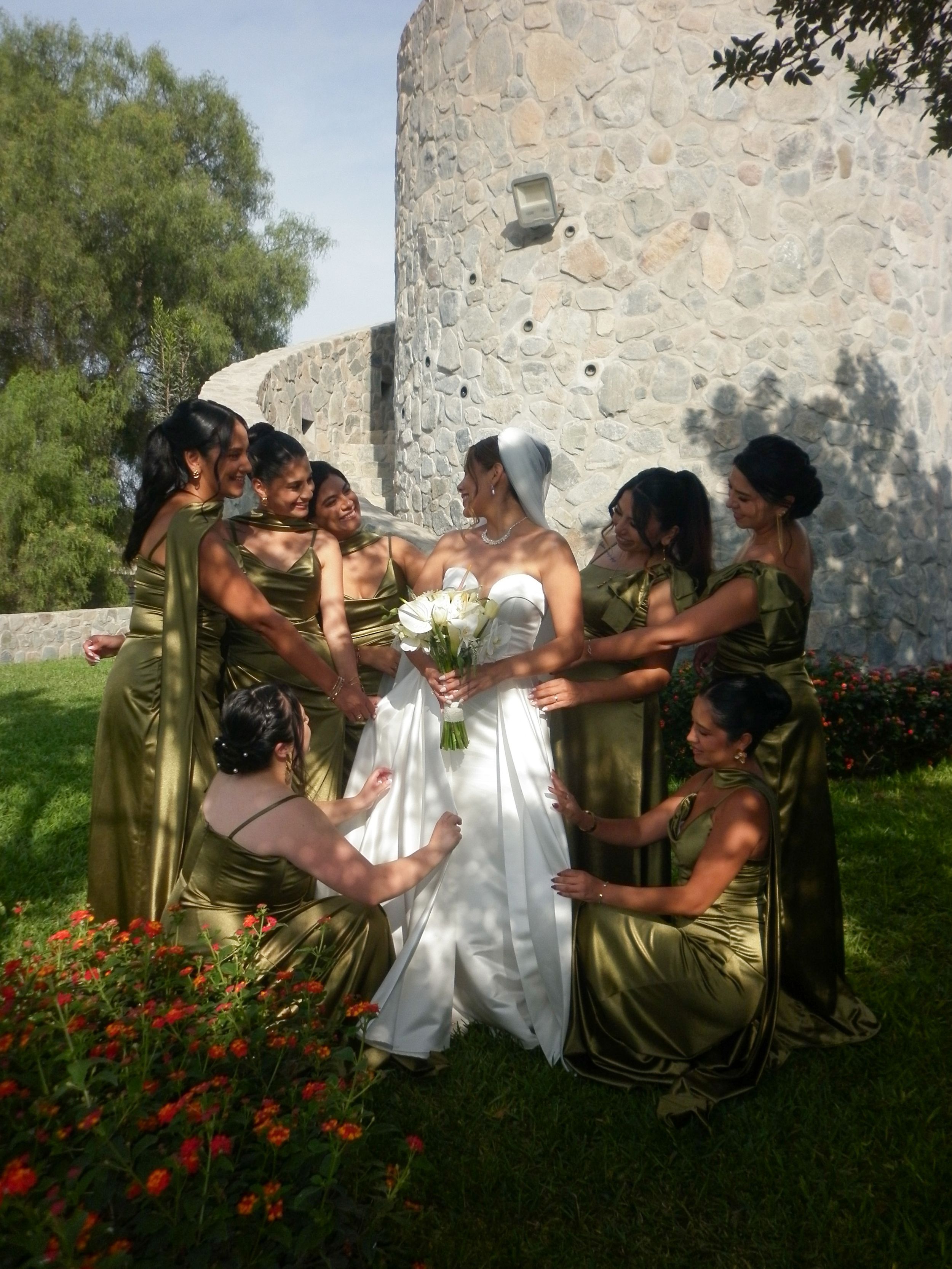 Bride in white gown surrounded by bridesmaids in gold dresses near a stone tower during outdoor wedding ceremony.