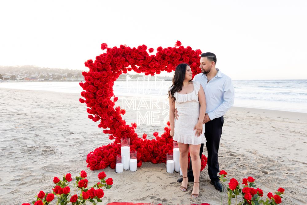 A couple stands together under a dramatic heart-shaped arch of red roses on a beach.