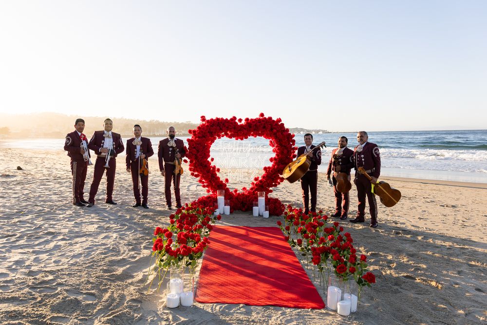 A romantic beach proposal setup with red rose petals forming a heart shape around a red carpet on sandy shore.
