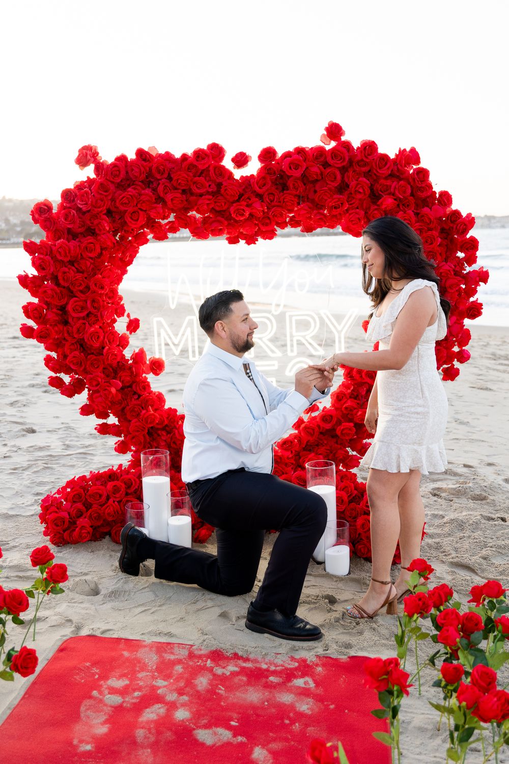 A romantic beach proposal setup with a large heart-shaped arch made of red roses on the sand at sunset.