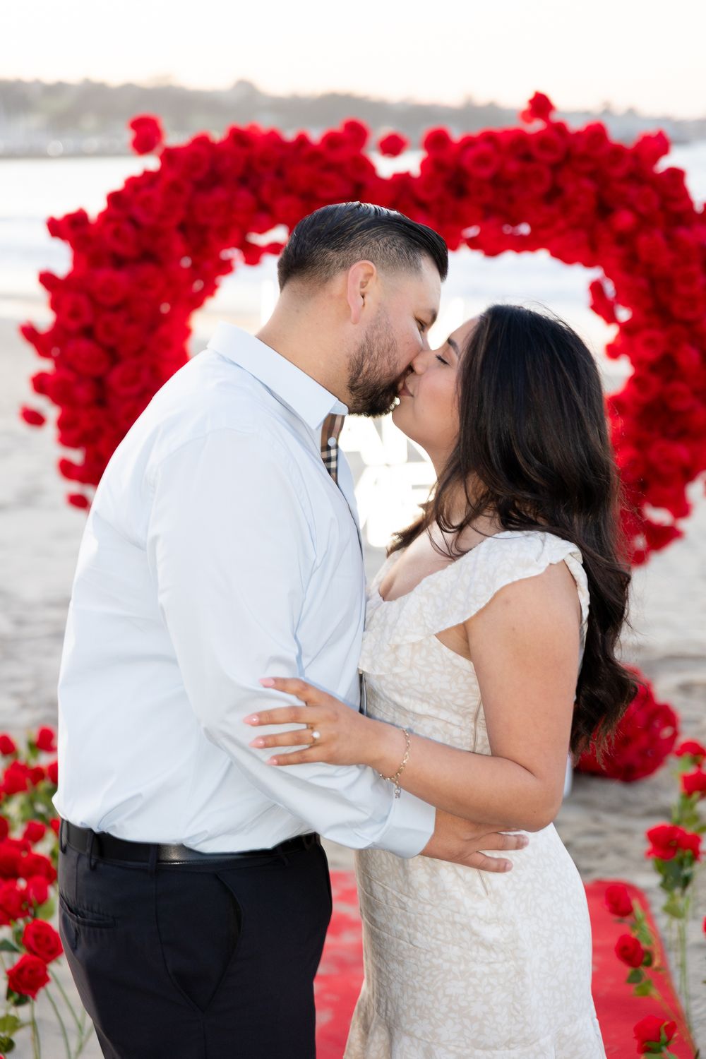 A romantic couple embraces by the water with vibrant red rose petals creating a stunning backdrop.