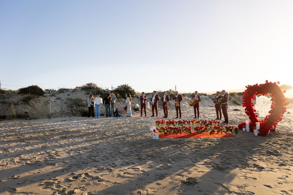 A line of people stand on a sandy beach at sunset with a bright red wreath glowing at the end of the line.