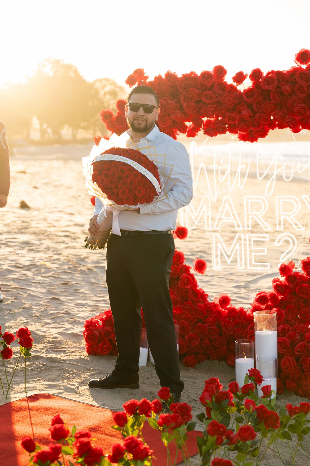 A romantic beach proposal setup with red roses scattered on a blanket at sunset.