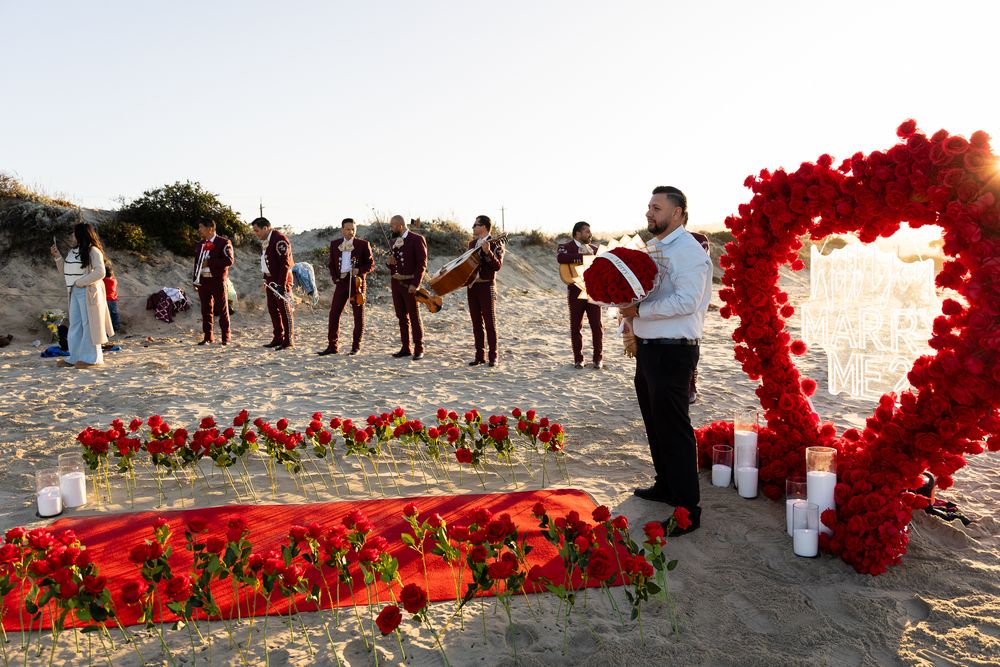 Red rose petals arranged in a heart shape on a sandy beach during sunset with people gathering nearby.