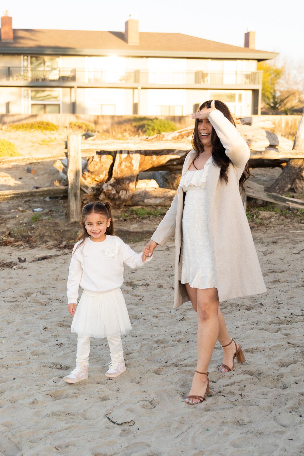 Two people in white outfits walking together on a sandy beach at sunset with buildings in the background.