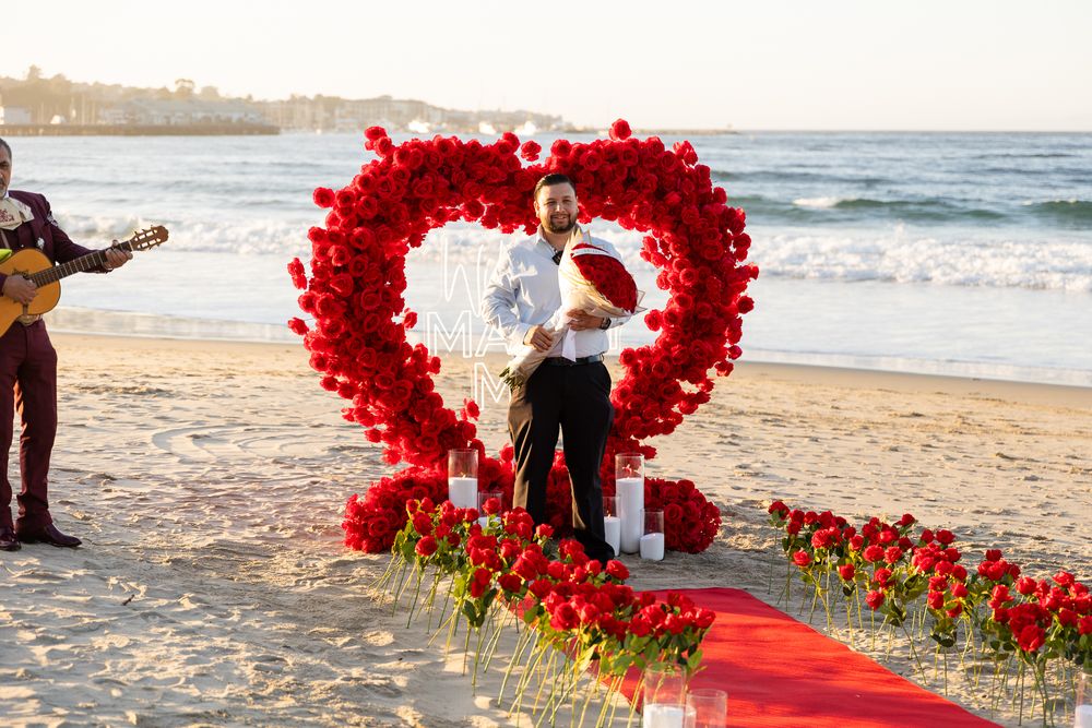 A romantic beach proposal setup features a heart-shaped arch made of red roses and a red carpet path leading to the ocean.