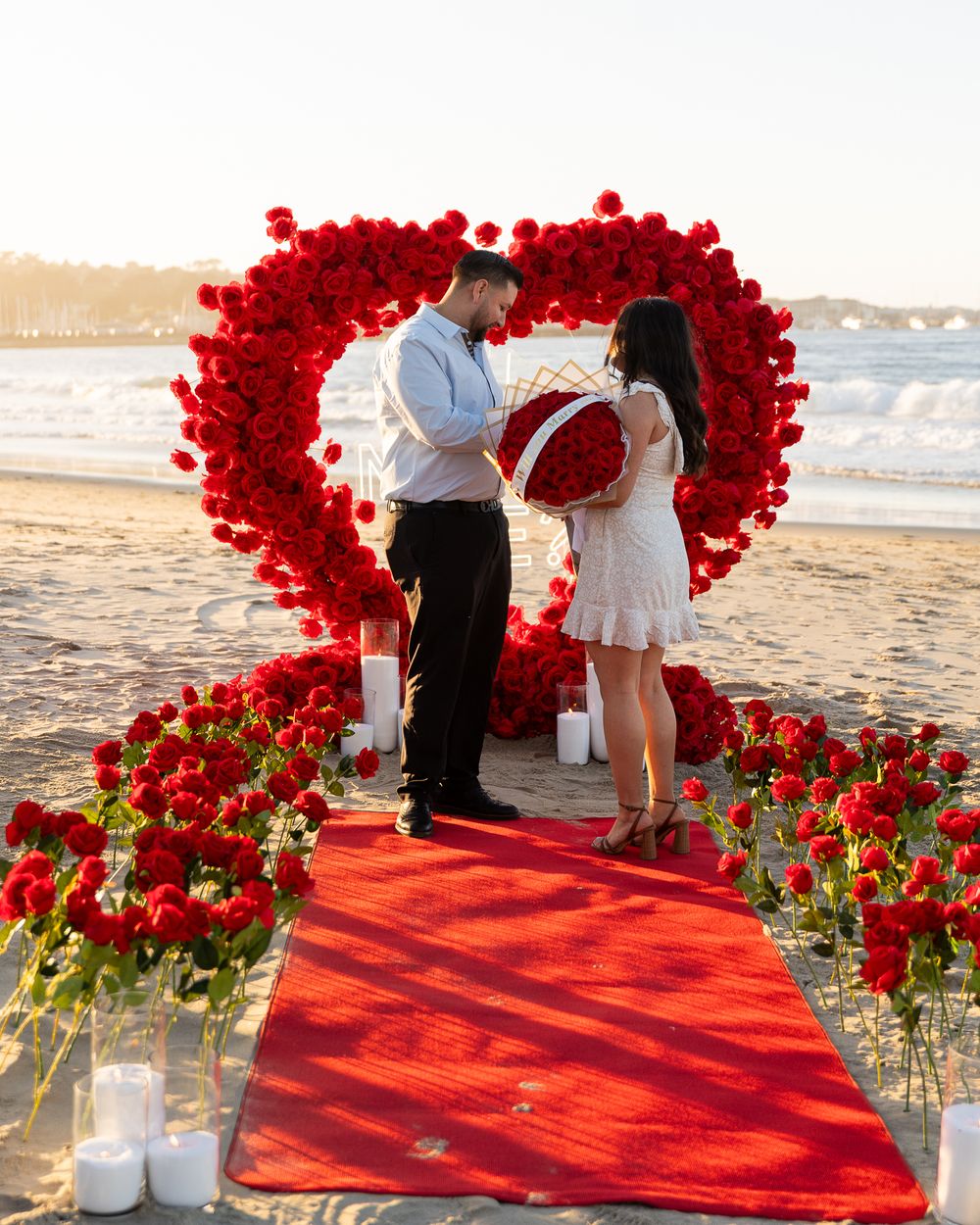 A romantic beach proposal setup with a heart-shaped arch made of red roses and a red carpet leading to the ocean.