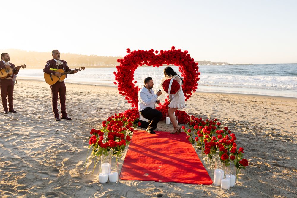 A beach proposal scene with musicians playing guitars as a couple stands under a red rose heart arch at sunset.