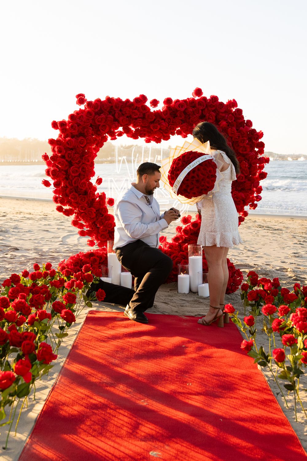 A romantic beach proposal setup with a heart-shaped arch made of red roses and a red carpet on the sand at sunset.