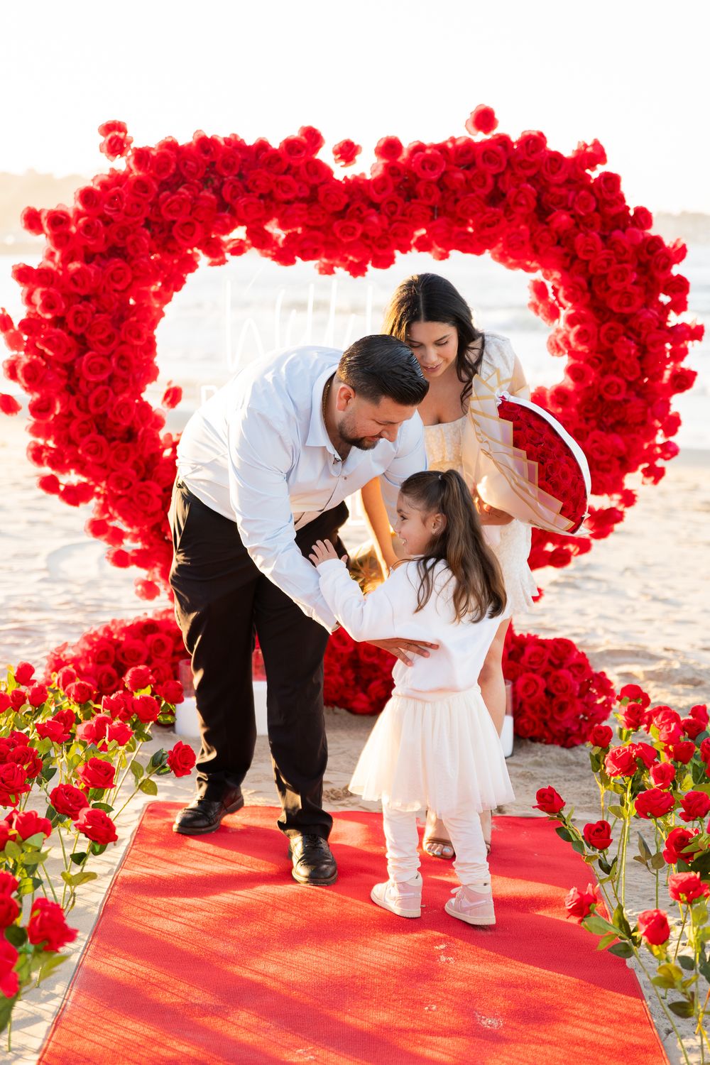 A family moment under a red rose heart arch on a beach with a red carpet path surrounded by flowers.