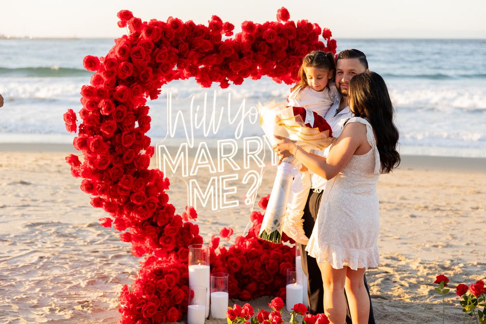 A romantic beach proposal setup with red rose petals and illuminated 'Marry Me' sign at sunset.