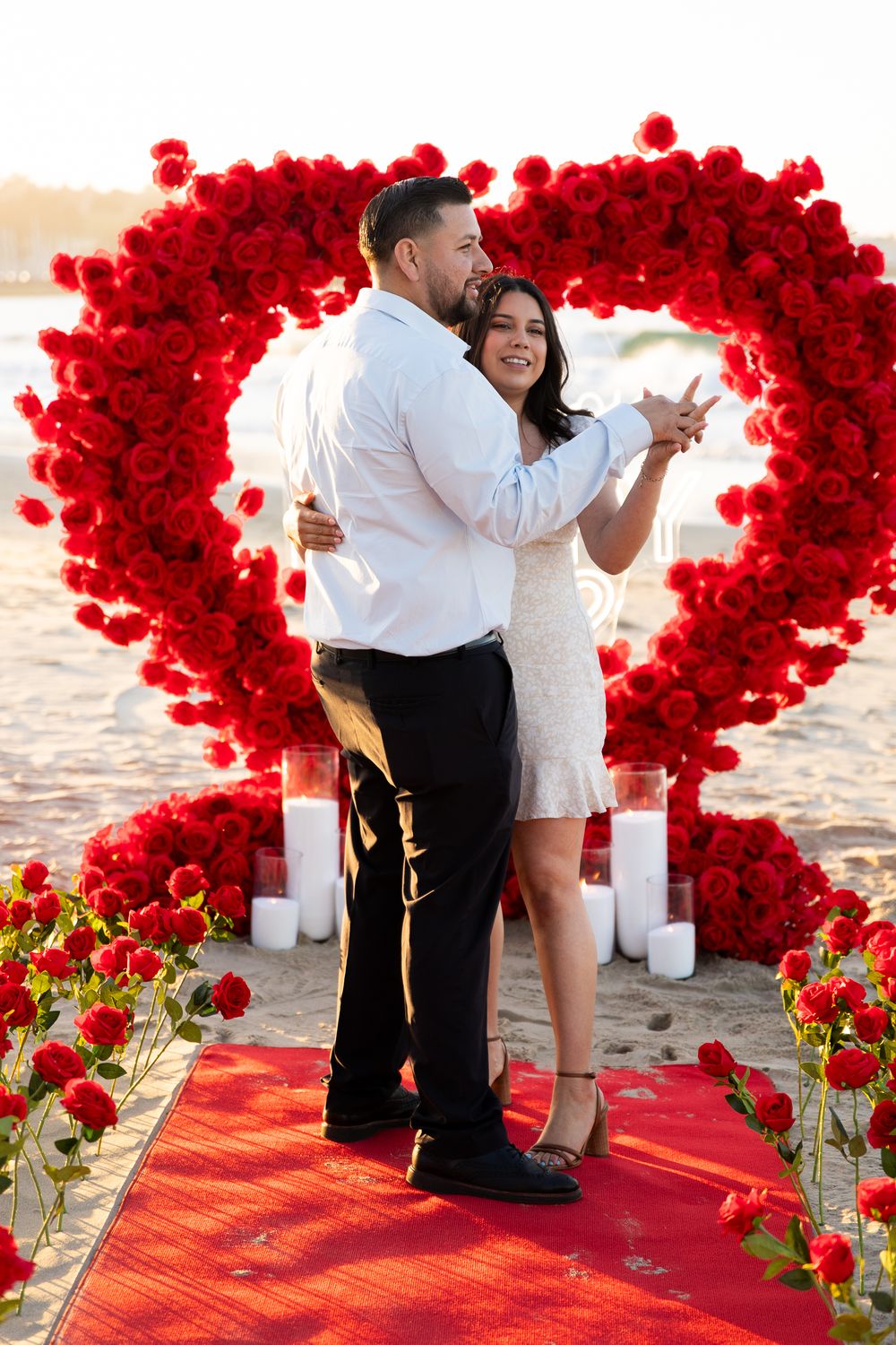 A romantic beach proposal setup with red rose petals forming heart shapes on a red carpet by the ocean.