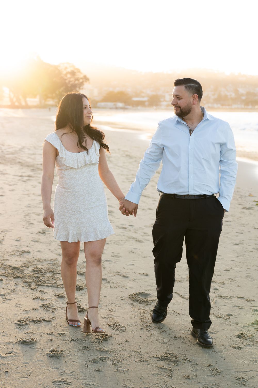 Couple walking hand in hand on a sandy beach during golden hour sunset, photographed from behind.