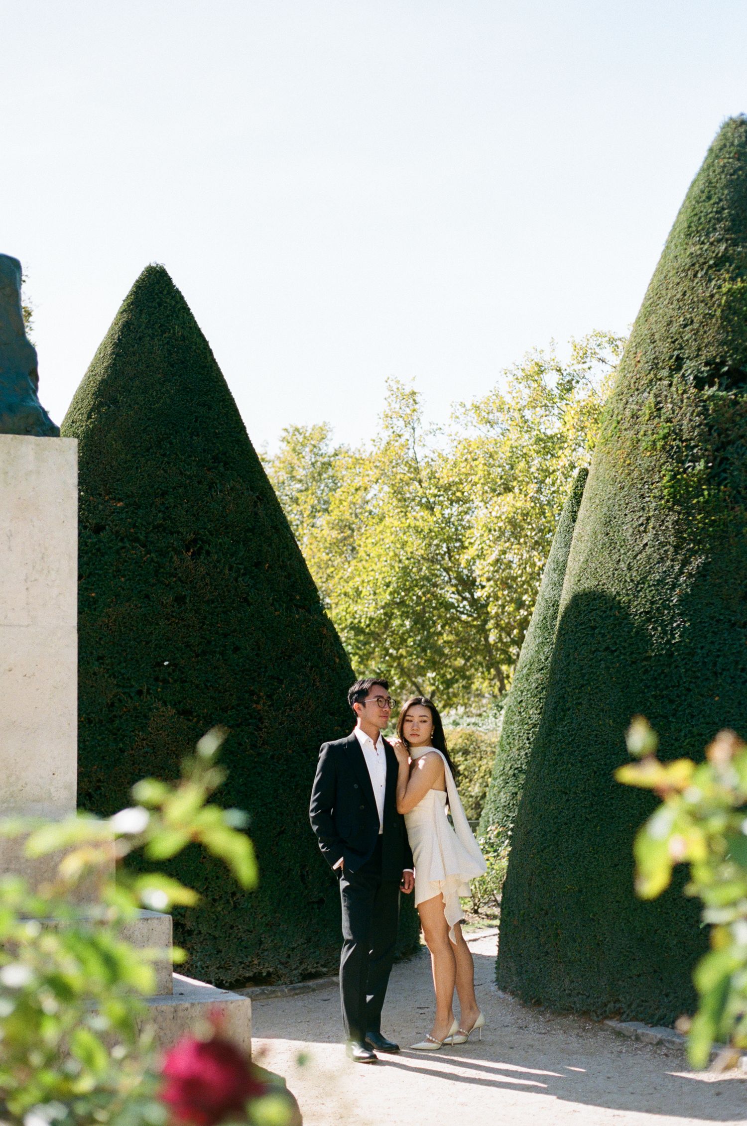Couple shares intimate moment between tall manicured hedges in elegant garden setting.