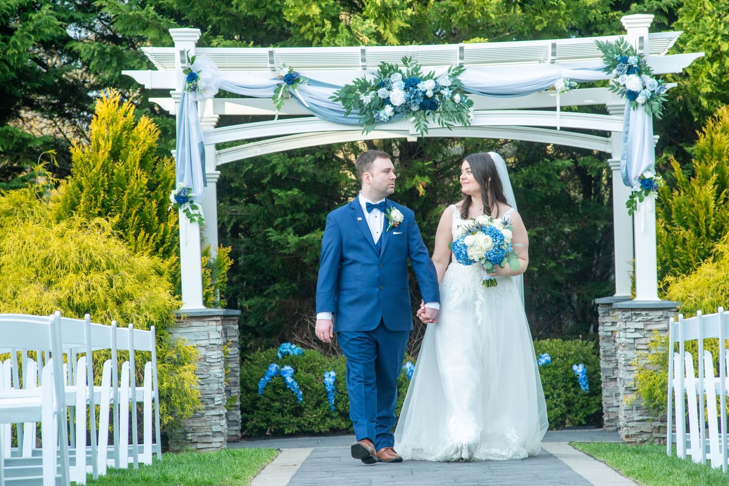 A couple stands under a white wedding arch decorated with blue and white flowers during an outdoor garden ceremony.