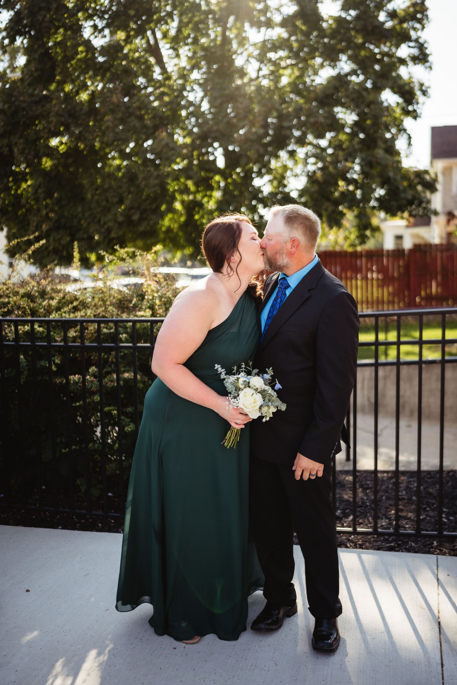 A couple shares a romantic kiss in formal attire with a green dress and black suit against a sunlit garden backdrop.
