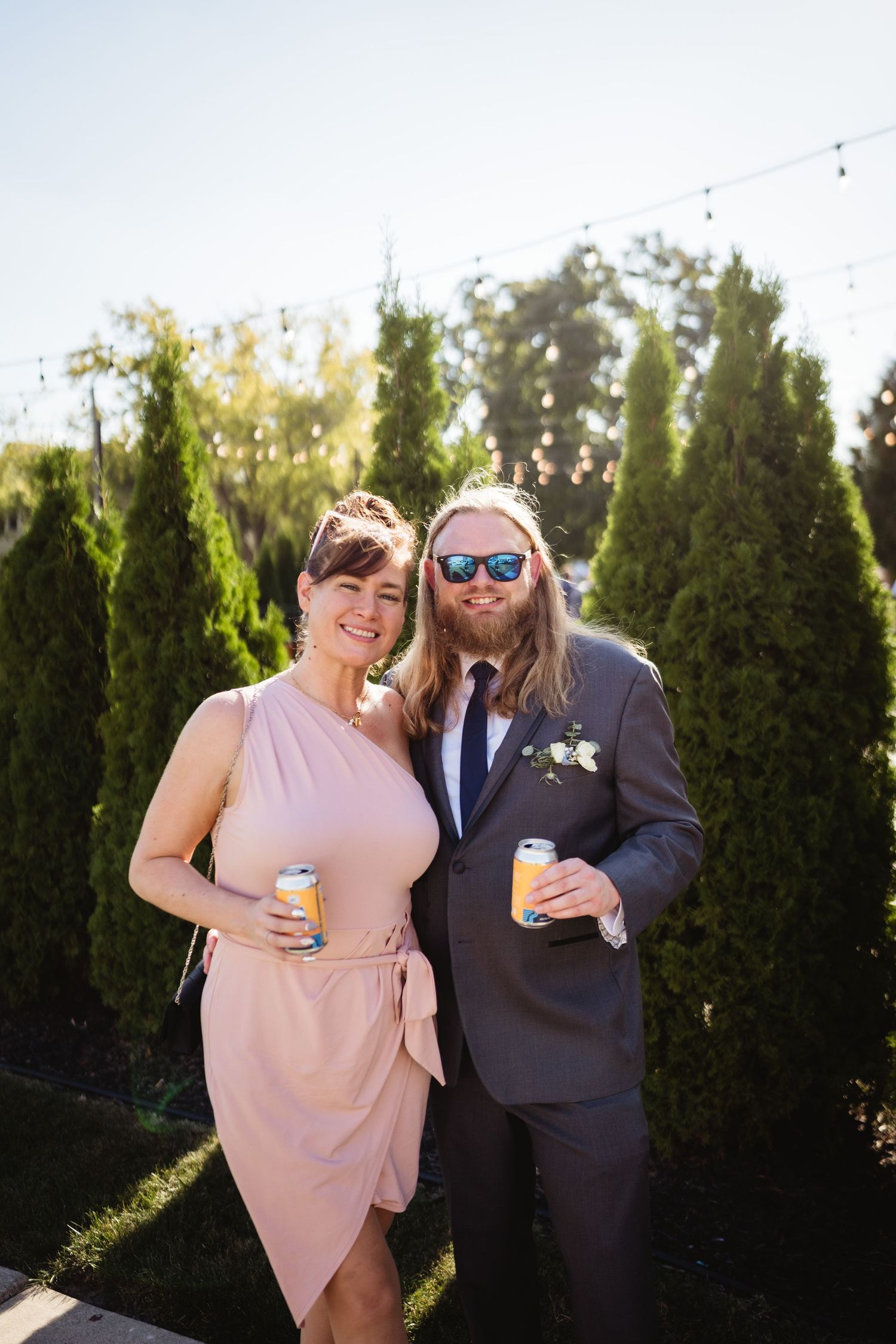 Two wedding guests in formal attire pose outdoors with drinks against a backdrop of evergreen trees.