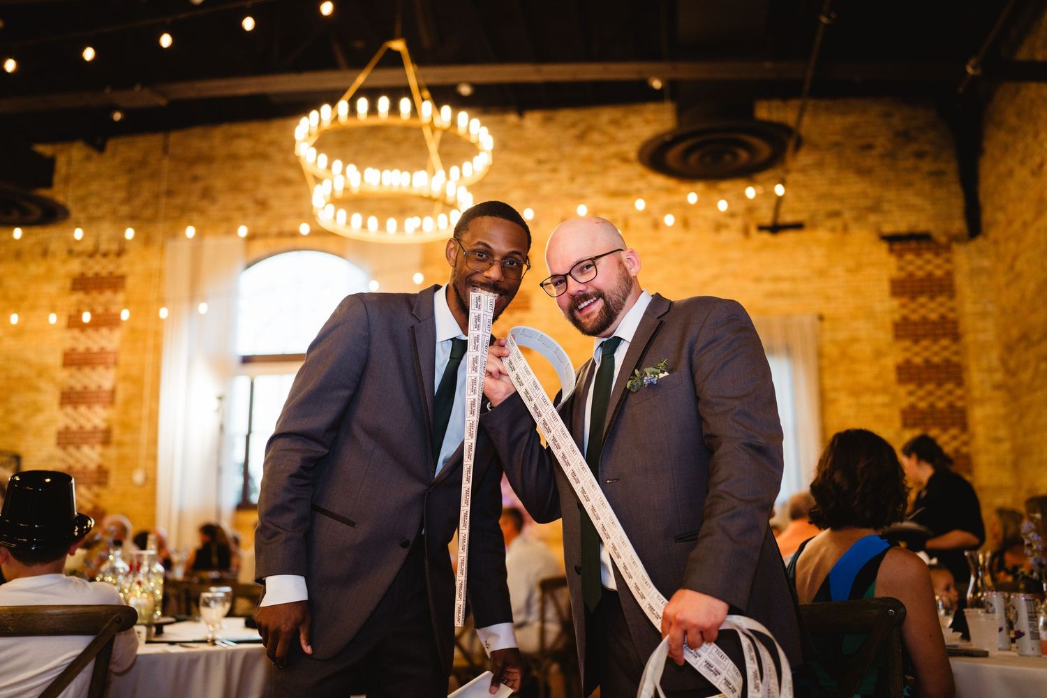 Wedding guests gather in an elegant rustic venue with exposed brick walls and glowing string lights overhead.