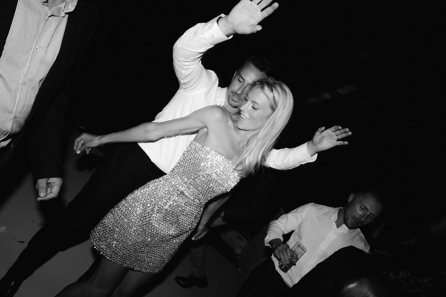 A person in a sparkly dress dances energetically with arms raised against a dark background.