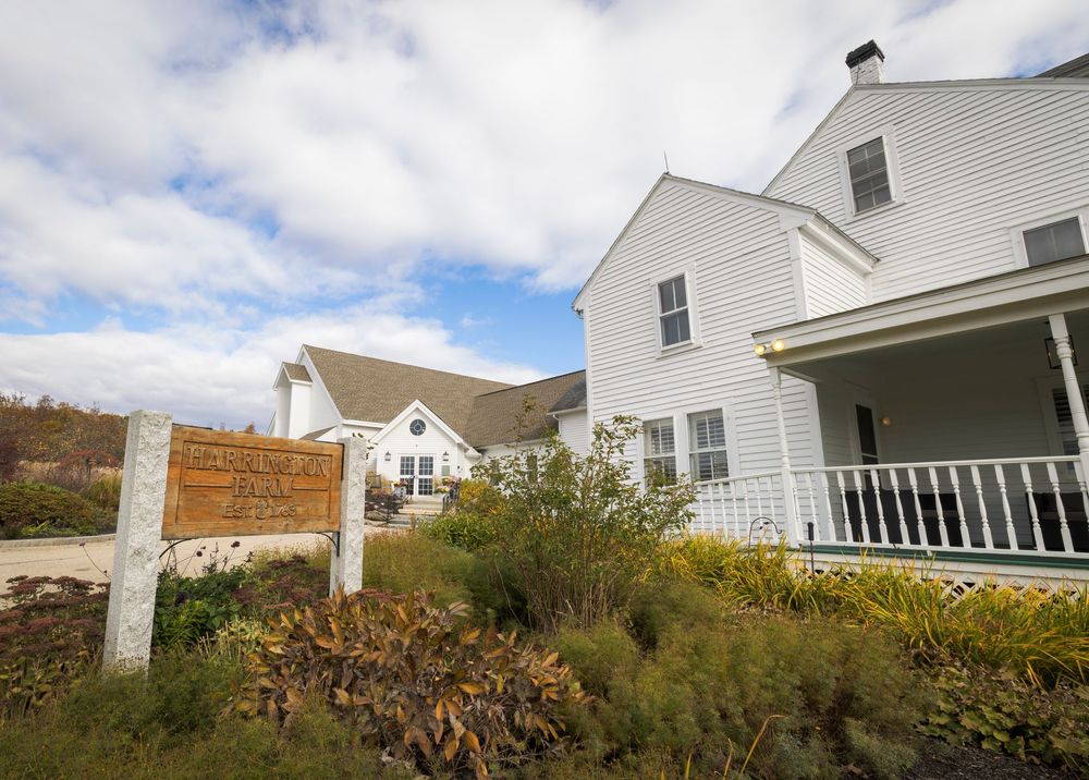 Row of white farmhouse-style buildings with porches set against blue sky and autumn foliage.