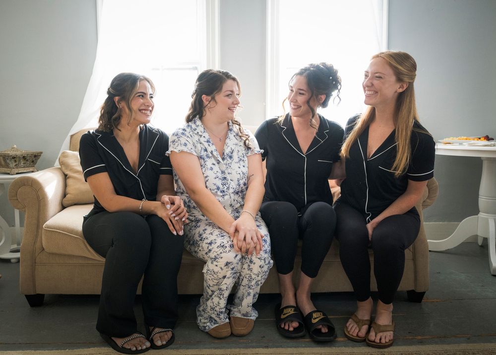 Group of wedding party members sit casually together on white furniture during pre-ceremony preparations.