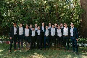 Large group of groomsmen in navy suits pose together outdoors under tree canopy for wedding photo.