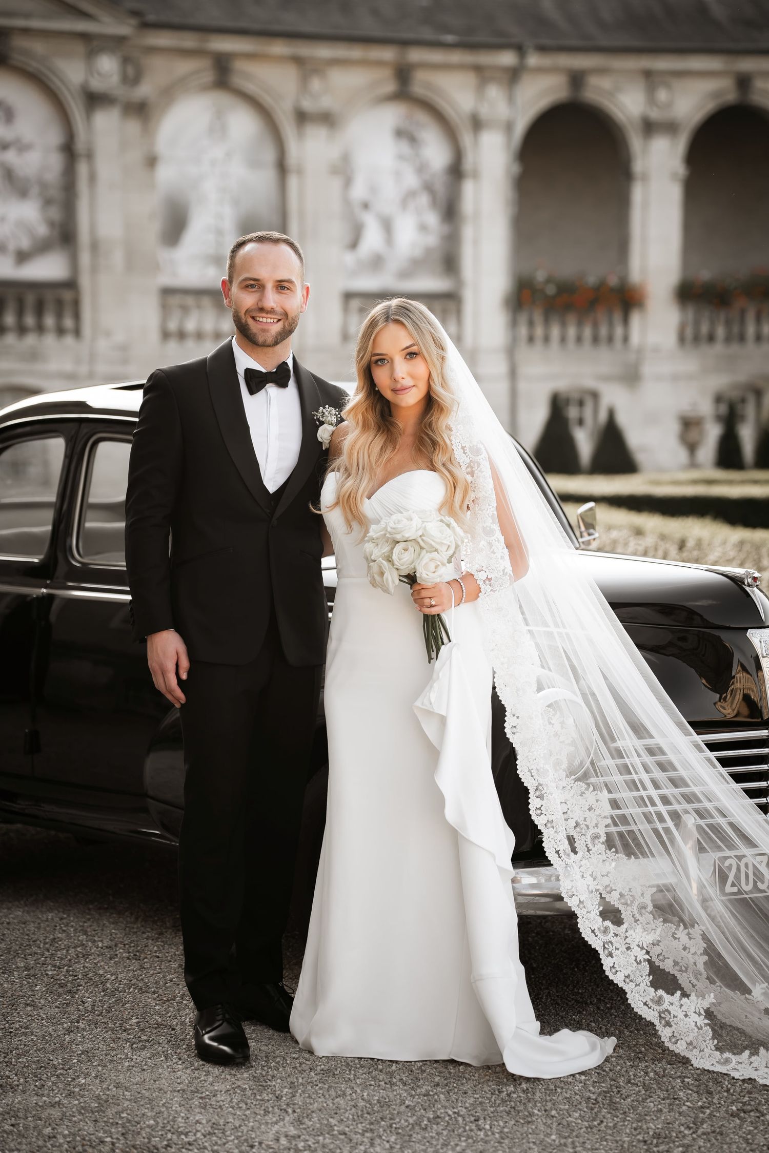 Elegant bride and groom posing beside a vintage black car in front of a grand European estate.