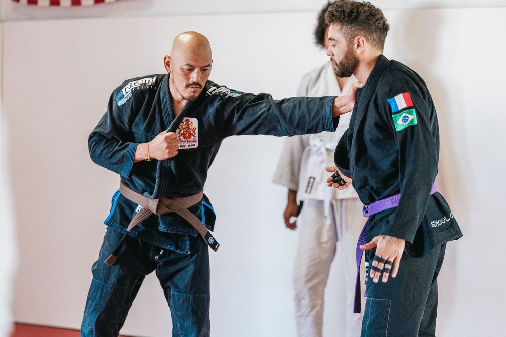 Two martial artists demonstrate a jiu-jitsu technique during training in a white-walled studio.