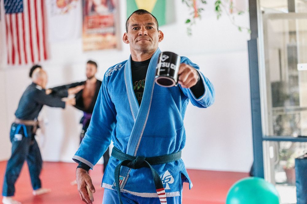 A martial artist in a blue gi holds up a beverage while training at a dojo with an American flag in the background.