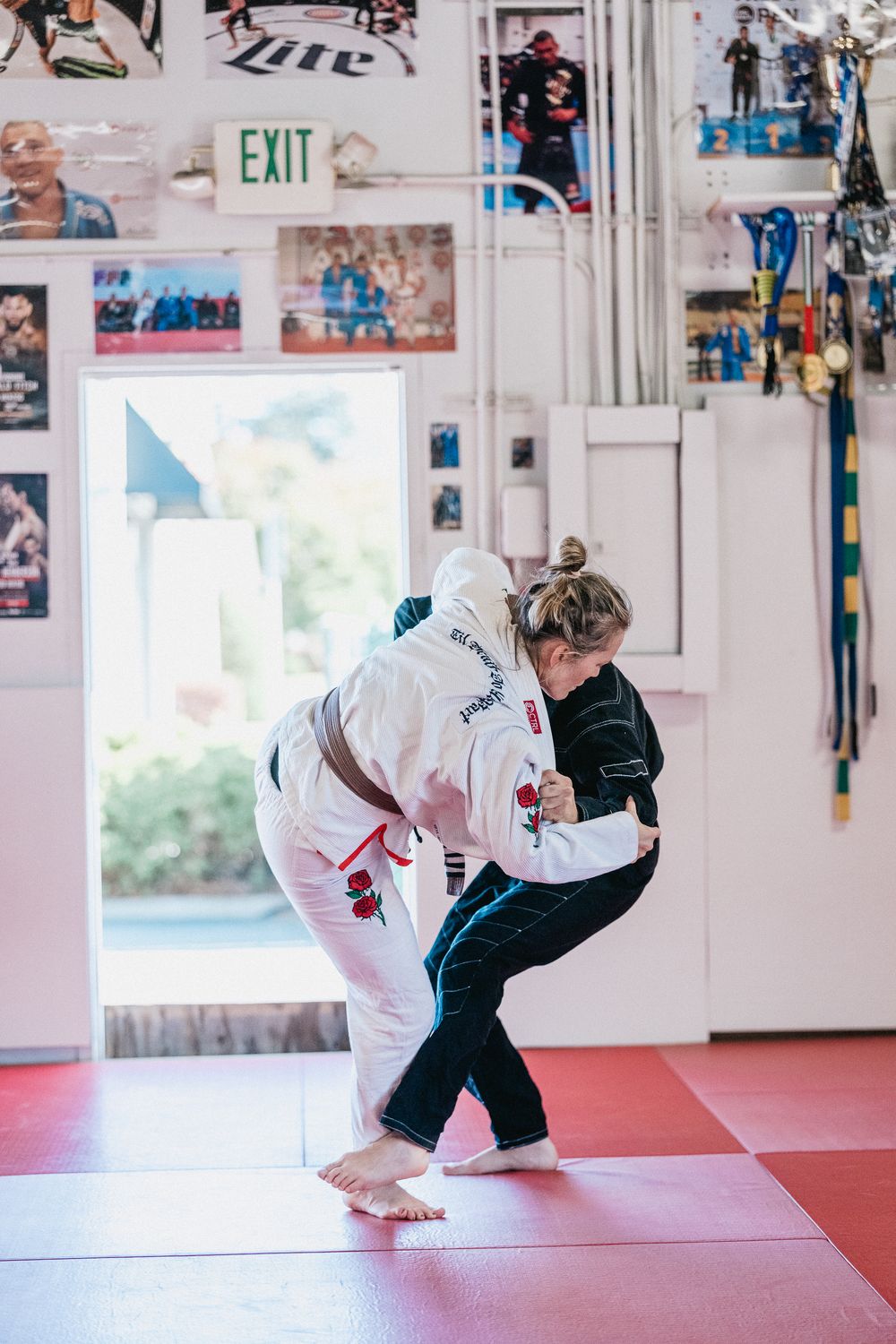 Two martial artists practicing grappling techniques on red training mats in a dojo with an exit sign visible.