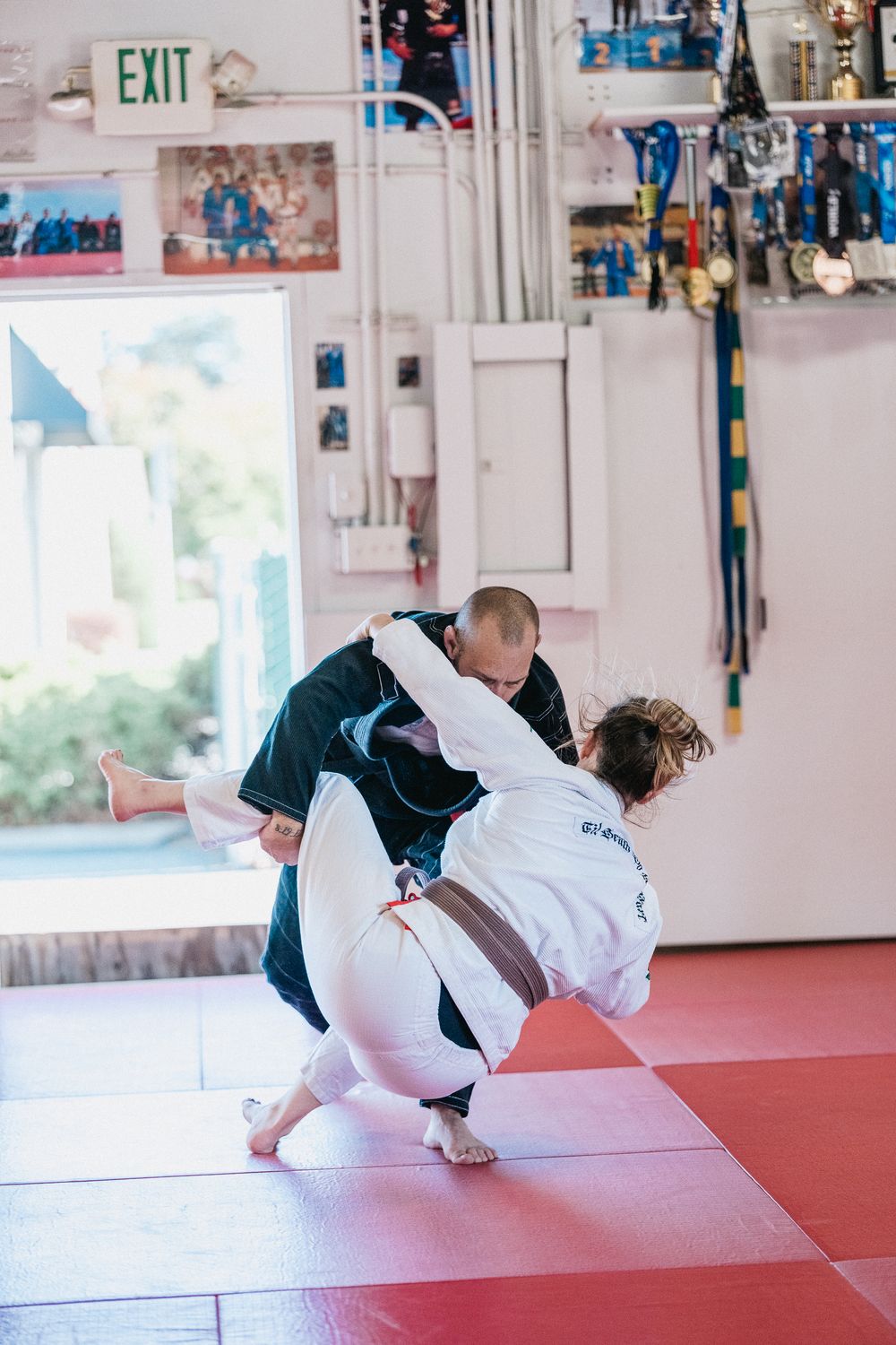 Students practicing martial arts rolls and breakfalls on red mats in a training studio.
