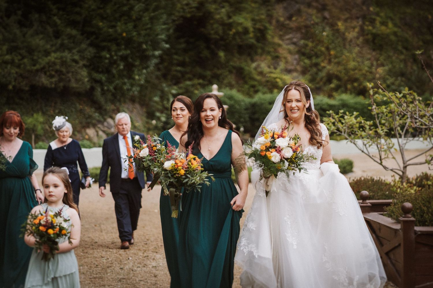 Wedding party members in emerald green dresses pose with bouquets outdoors.