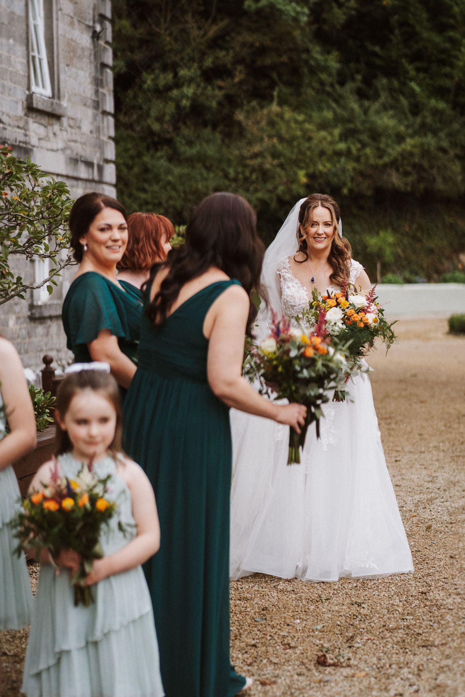 Wedding party in emerald green dresses and white gown gather outdoors for photos near stone building.