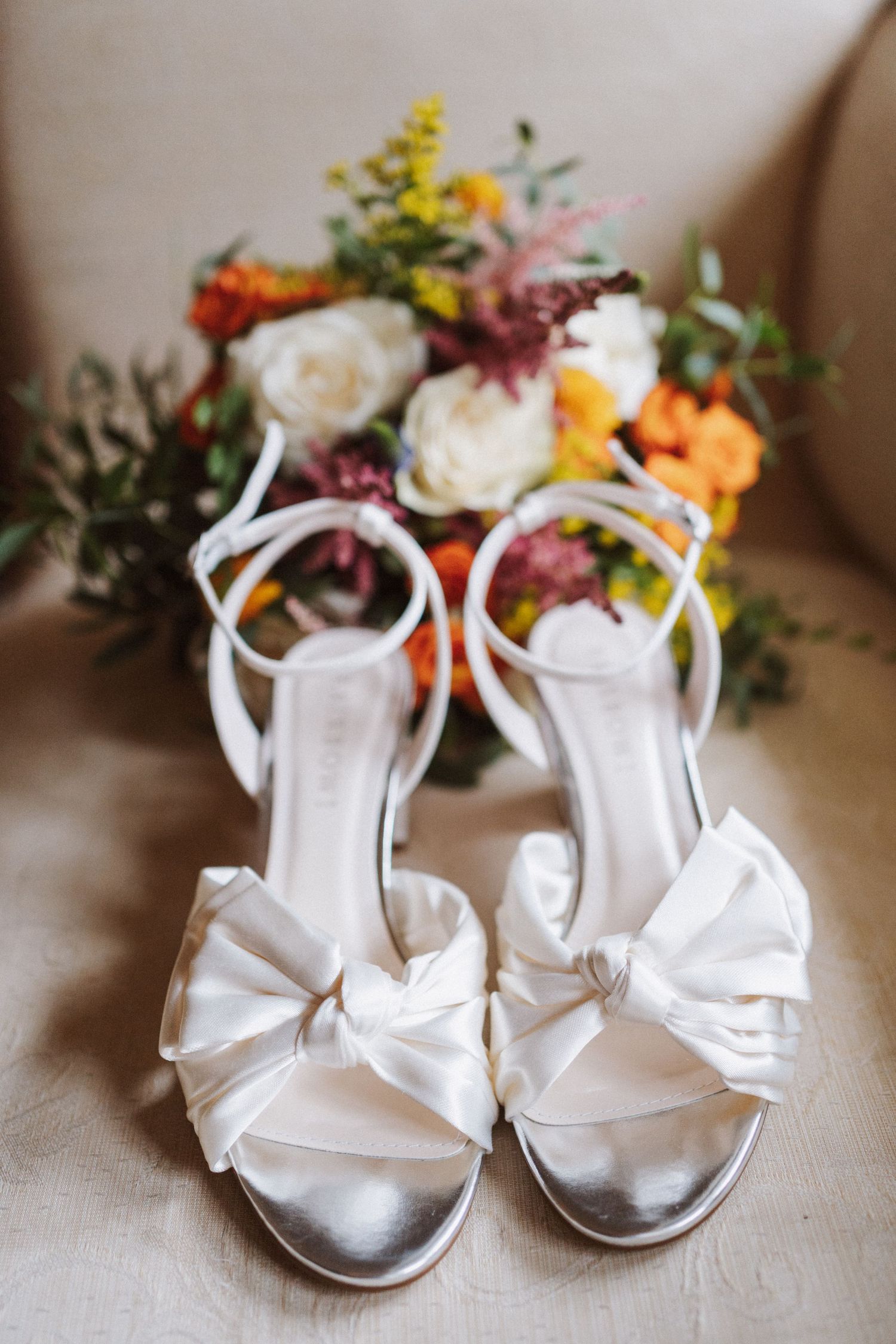 White satin wedding shoes with ankle straps and floral bouquet decorations in the background.