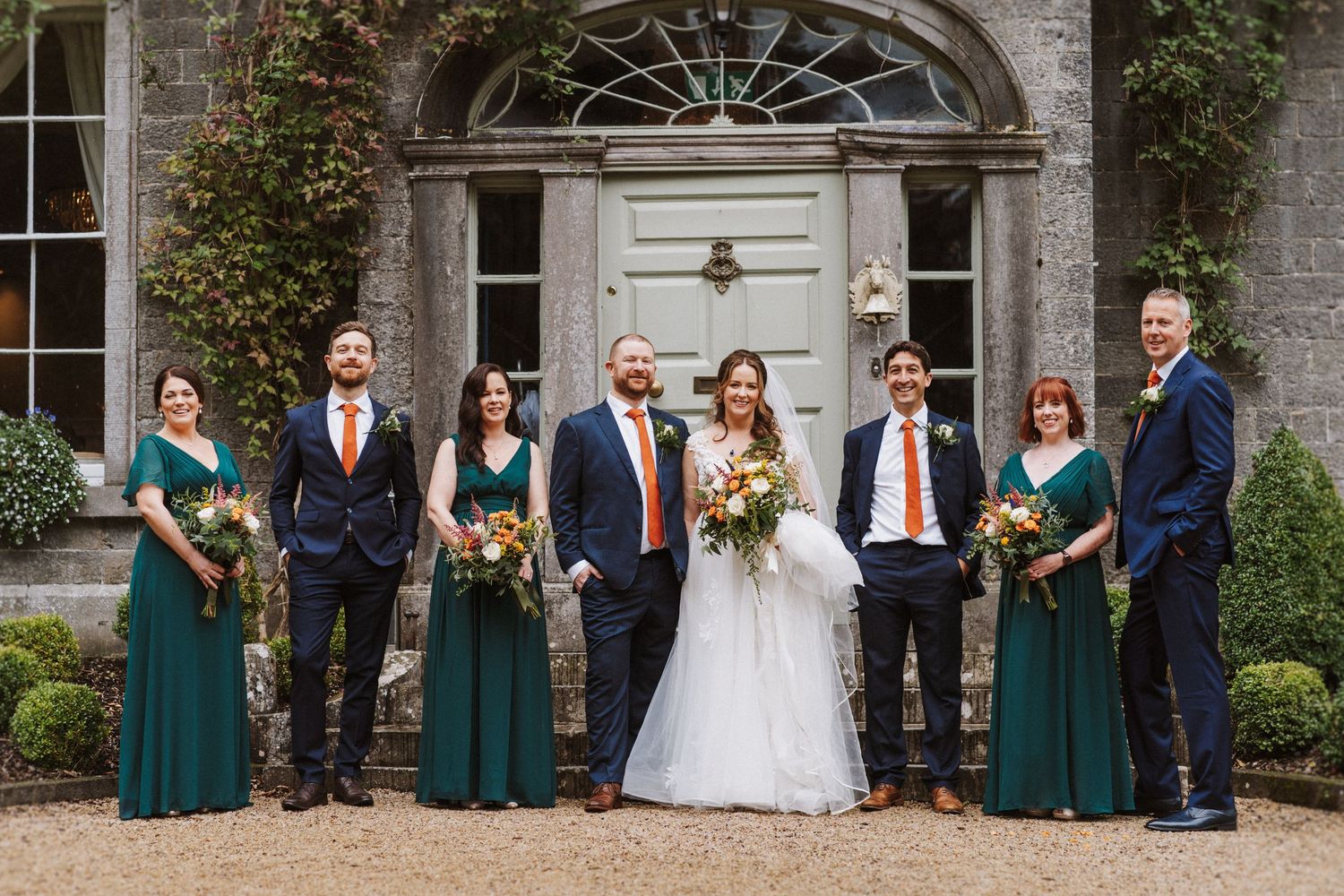 Wedding party poses in front of elegant stone mansion with emerald green bridesmaid dresses and autumn florals.