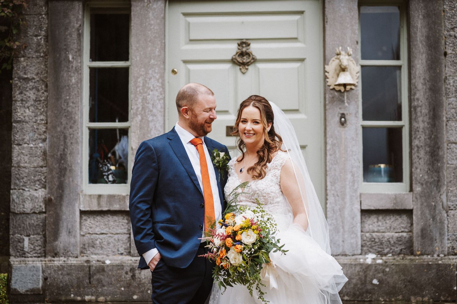 A happy couple shares a joyful moment during their wedding ceremony outside a stone building with yellow flowers.