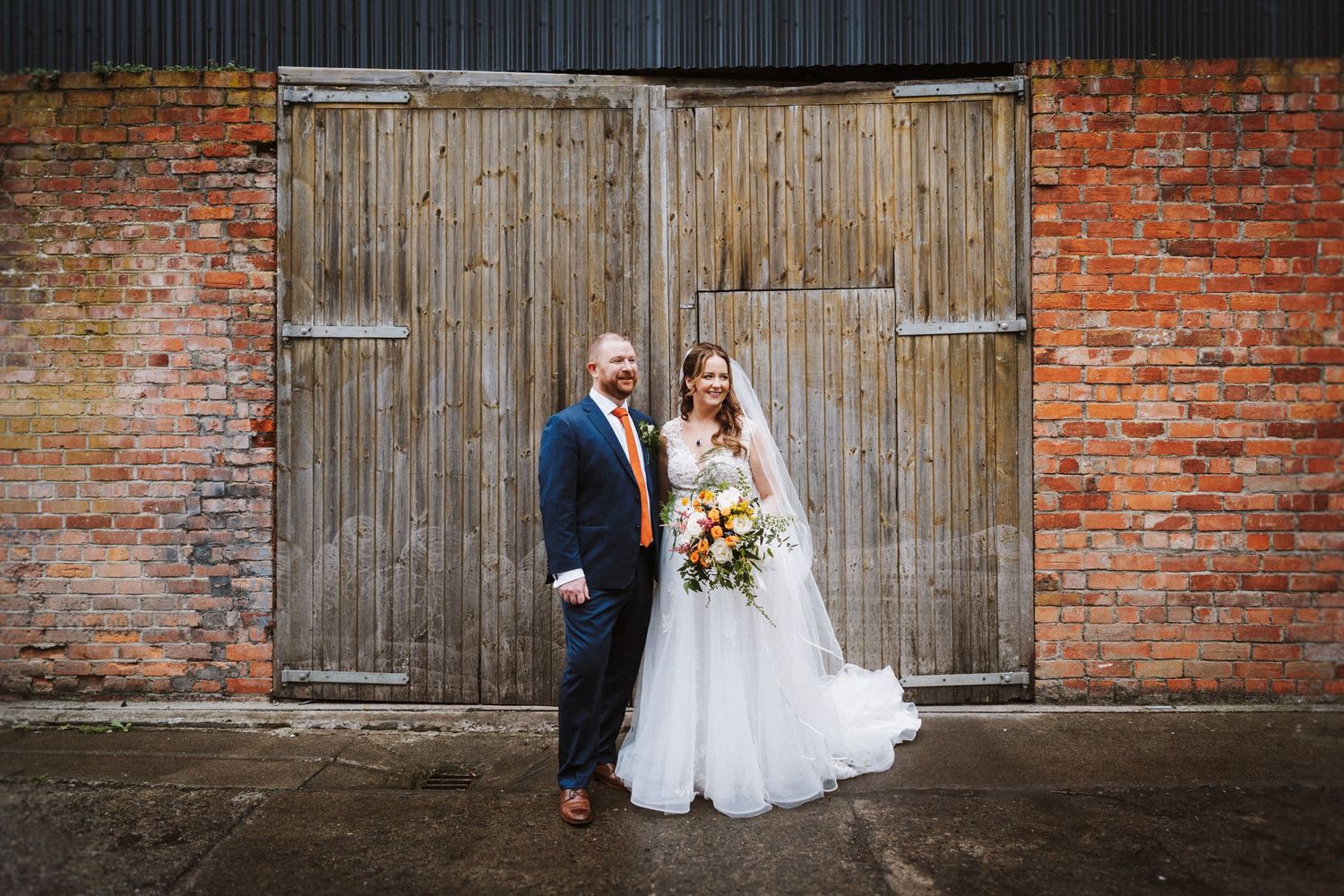 Wedding couple and party pose against rustic brick wall with wooden barn doors.