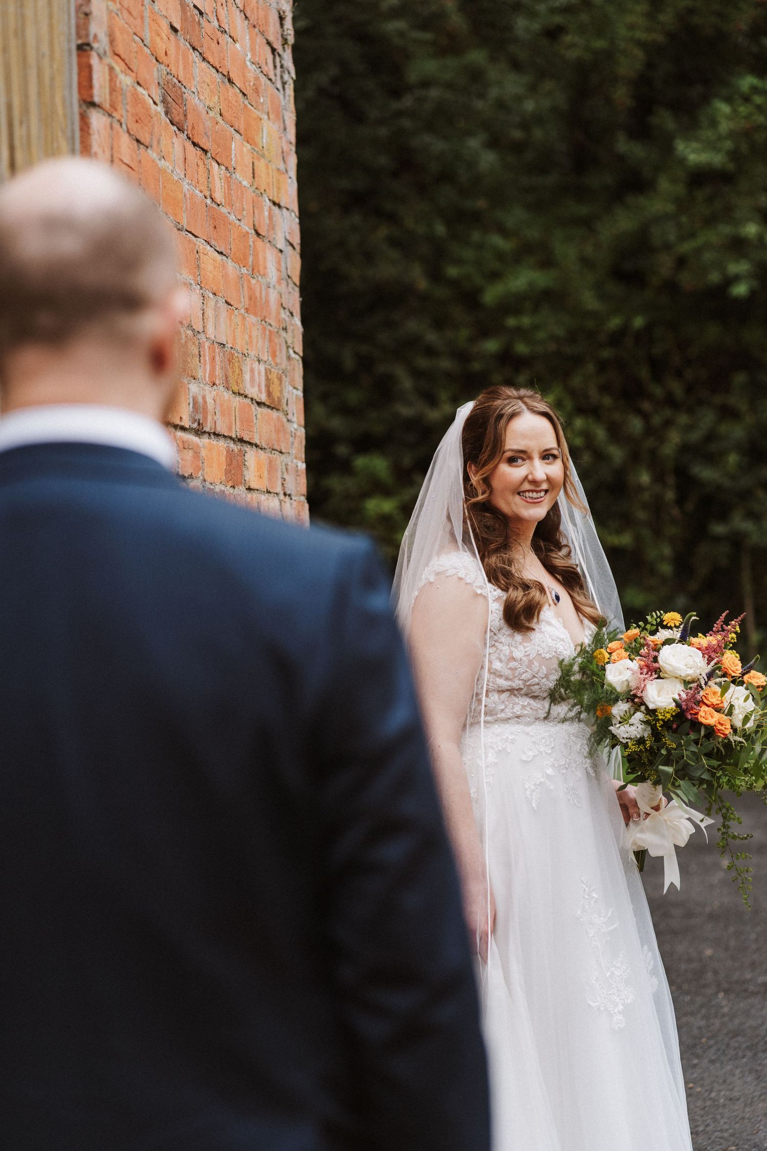 A bride in a white lace wedding dress holds a vibrant autumn bouquet against a brick wall backdrop.