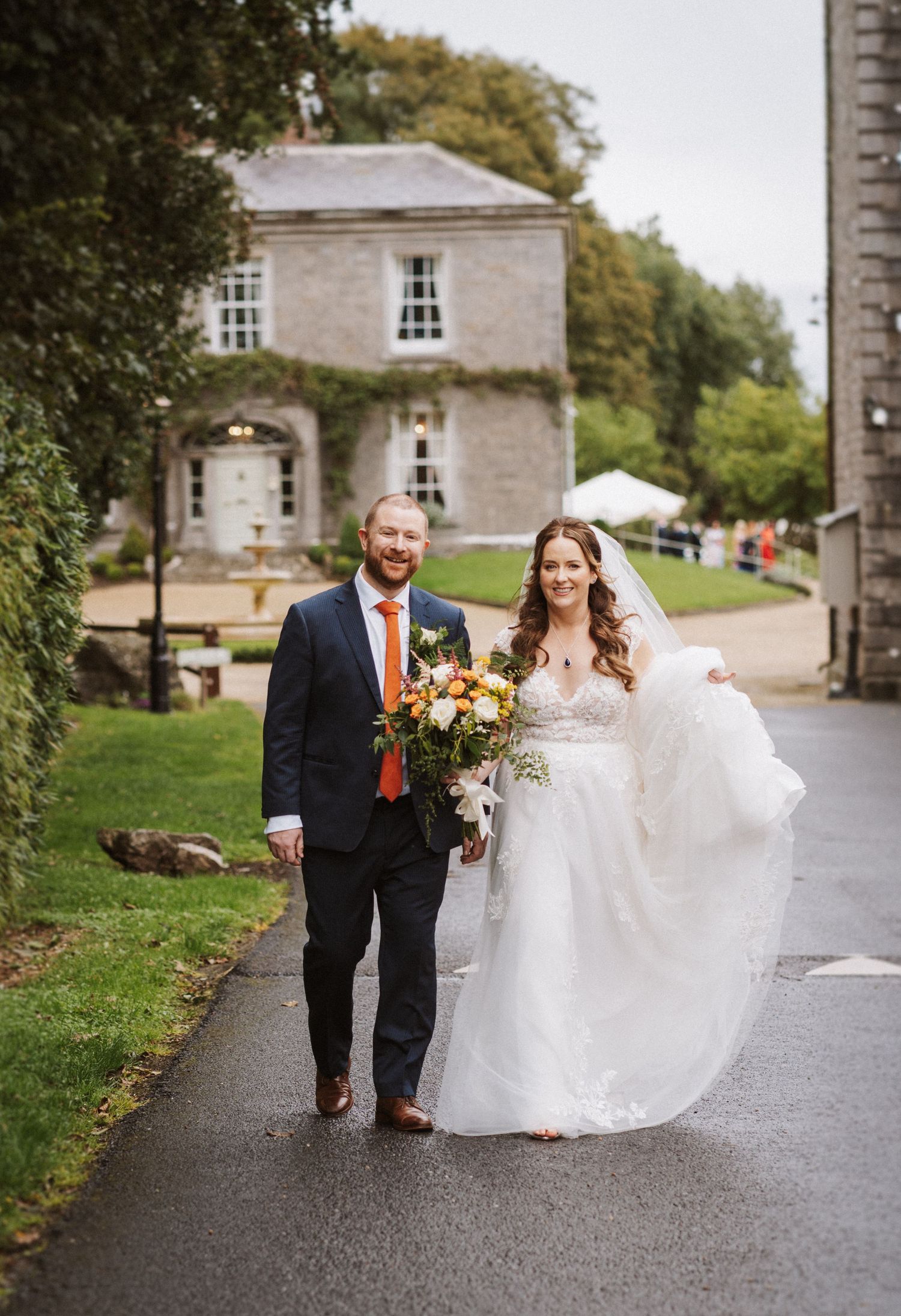 A couple walking together on a stone path surrounded by autumn trees, with the bride's white dress flowing in the breeze.