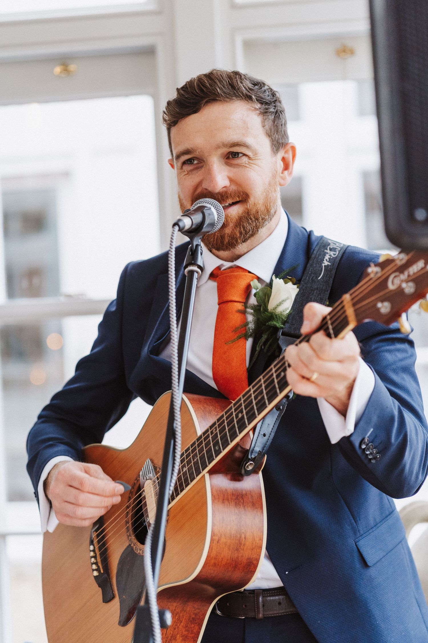 A musician in a navy suit and orange tie performs with an acoustic guitar during a wedding reception.