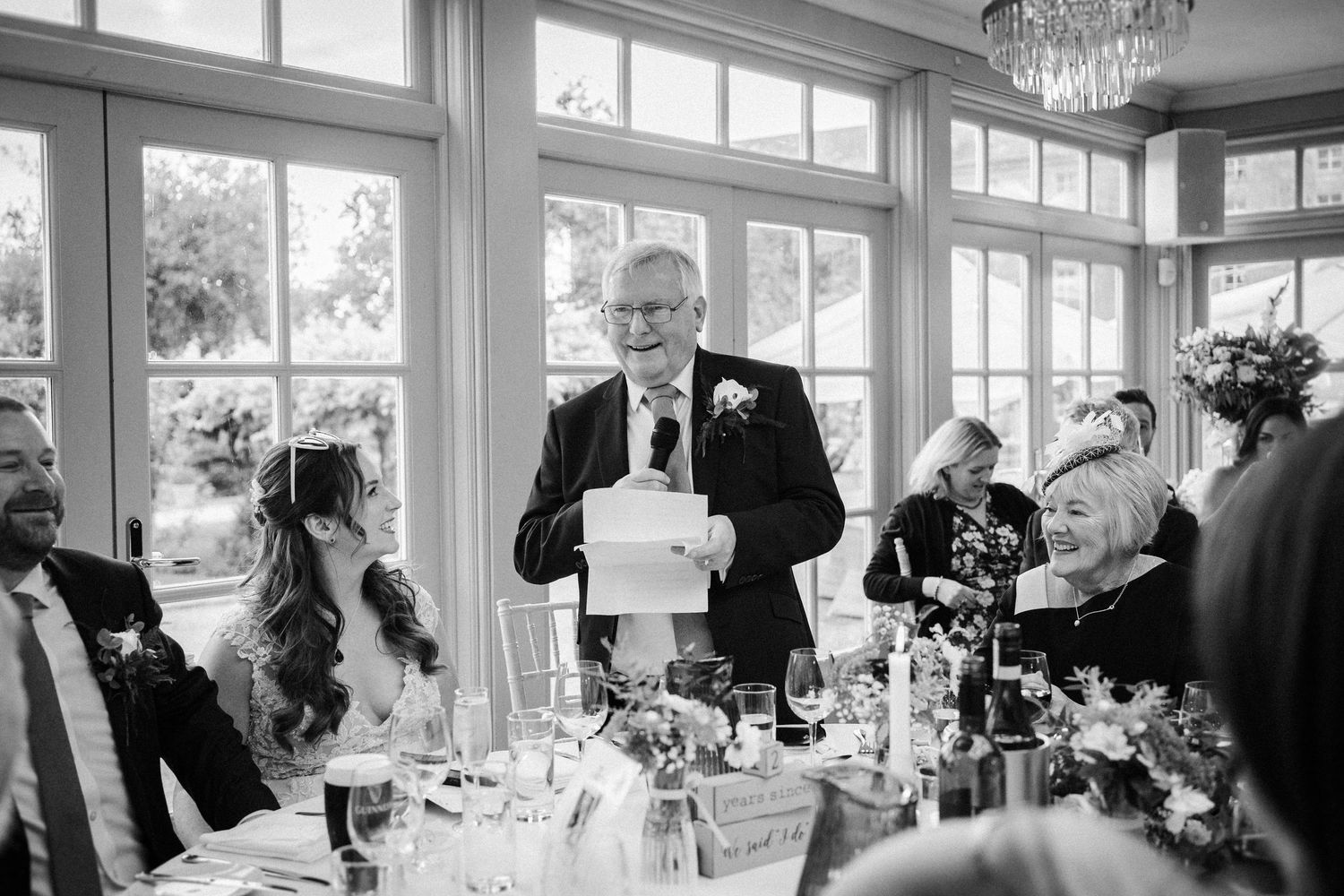 A person delivers a wedding speech at an elegant reception venue with large windows and crystal chandeliers.