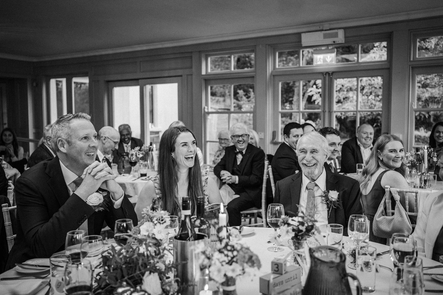 Black and white photo of guests laughing and enjoying themselves at an elegant dinner reception with wine glasses and flowers.