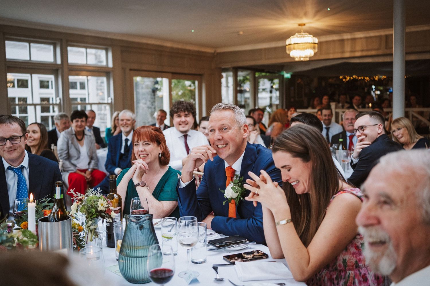 Wedding guests share laughter and conversation around elegant dining tables at an indoor reception venue.