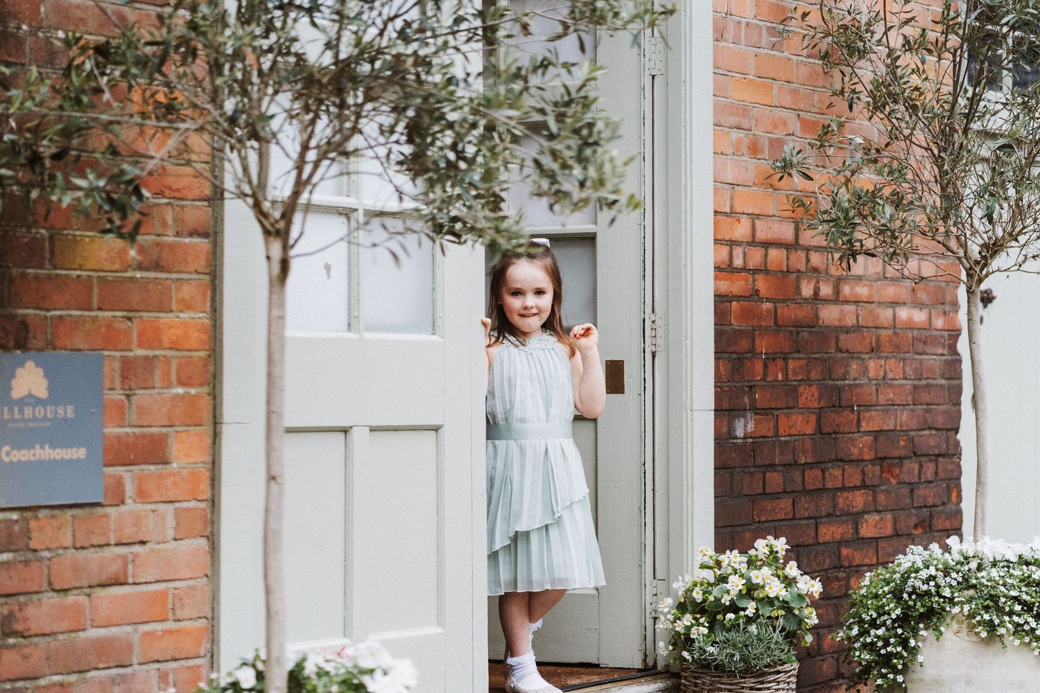 A person in a light blue dress stands in front of a white door and brick wall with climbing vines.