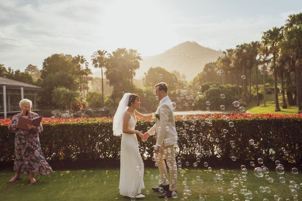 A couple stands together during a sunset wedding ceremony in a garden with golden sunlight filtering through trees.